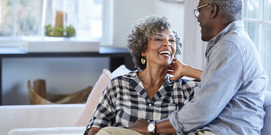 Una hermosa pareja negra senior con camisas largas se sienta mirándose en un sofá en una bonita sala de estar.