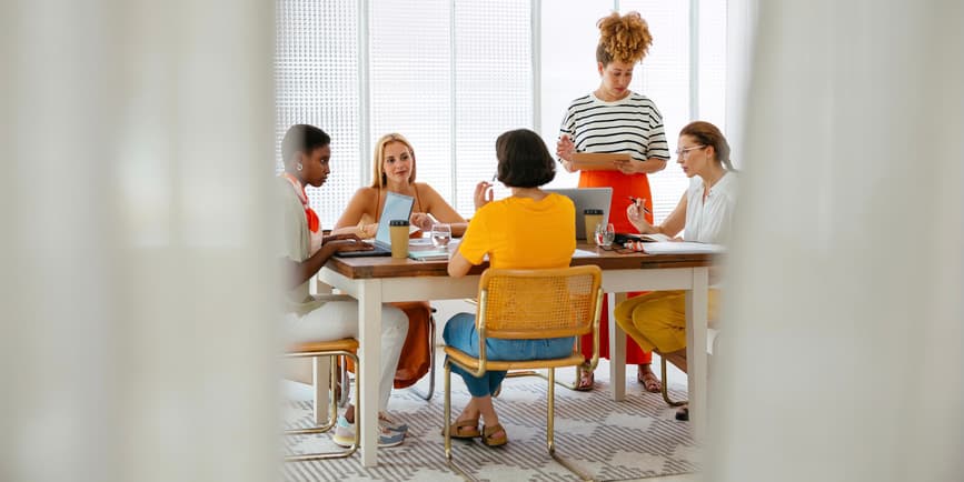 Grupo de compañeras vestidas de manera casual sentadas a la mesa con un portátil y discutiendo los detalles del proyecto mientras trabajan juntas en un espacio de trabajo conjunto