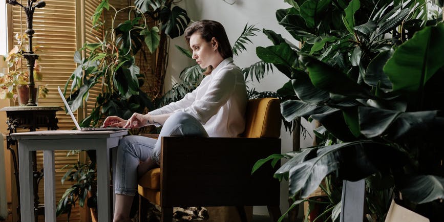 Side view of young woman in white shirt sitting in yellow chair. She bending leg and typing notes on laptop on grey table in studio with tropical plants, looking at screen