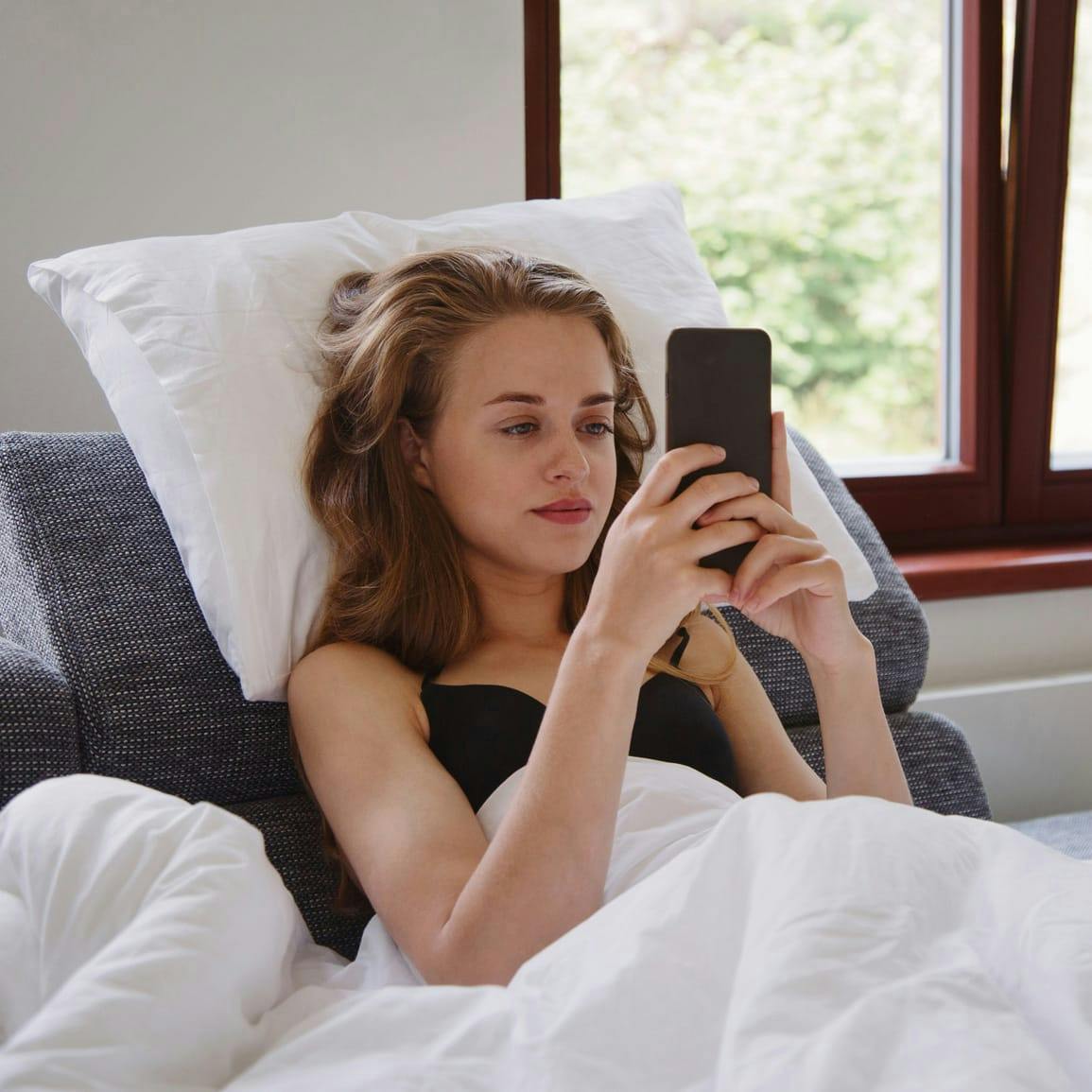 Mujer sentada en la cama, cubierta de sábanas, con las dos manos en el teléfono