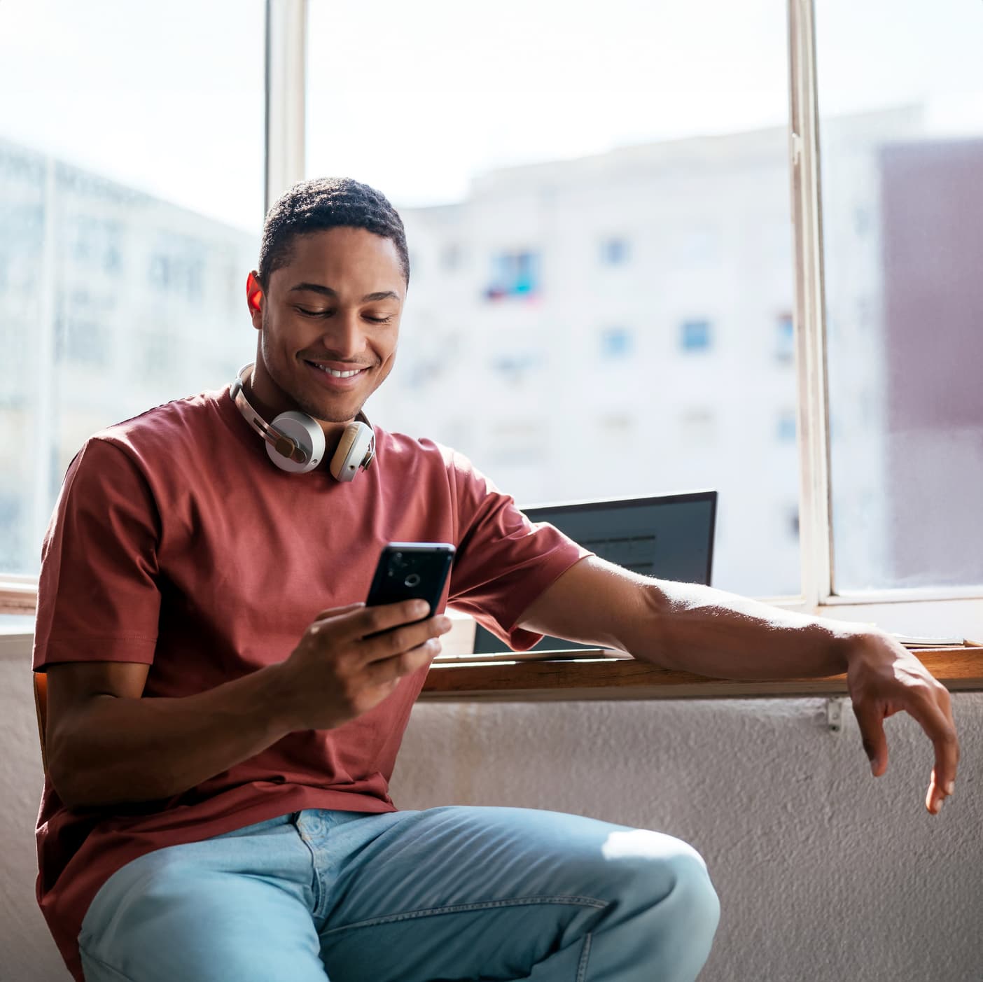 Hombre negro mirando su teléfono mientras está en un escritorio junto a una ventana, sonriendo