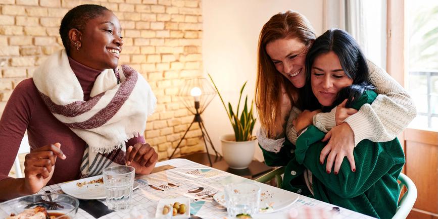 Amigas vestidas de invierno se reúnen en una mesa, sonriendo y abrazándose.