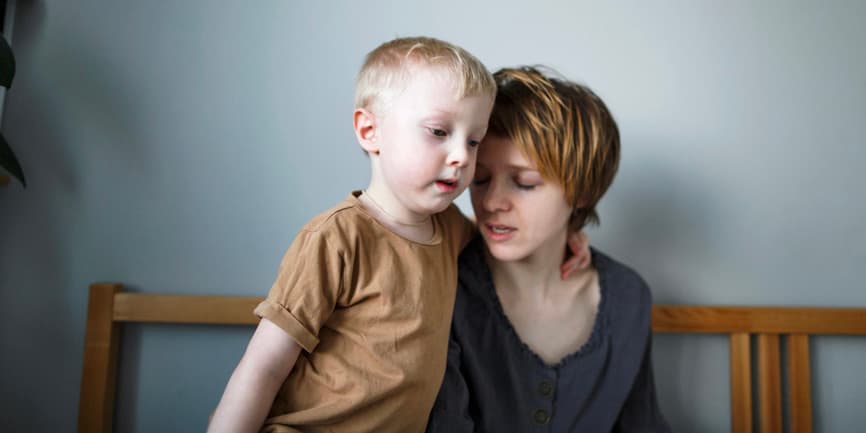 Una joven madre blanca con el pelo corto y rojo se sienta en la cama con aspecto cansado mientras habla con su joven hijo rubio que la abraza suavemente.