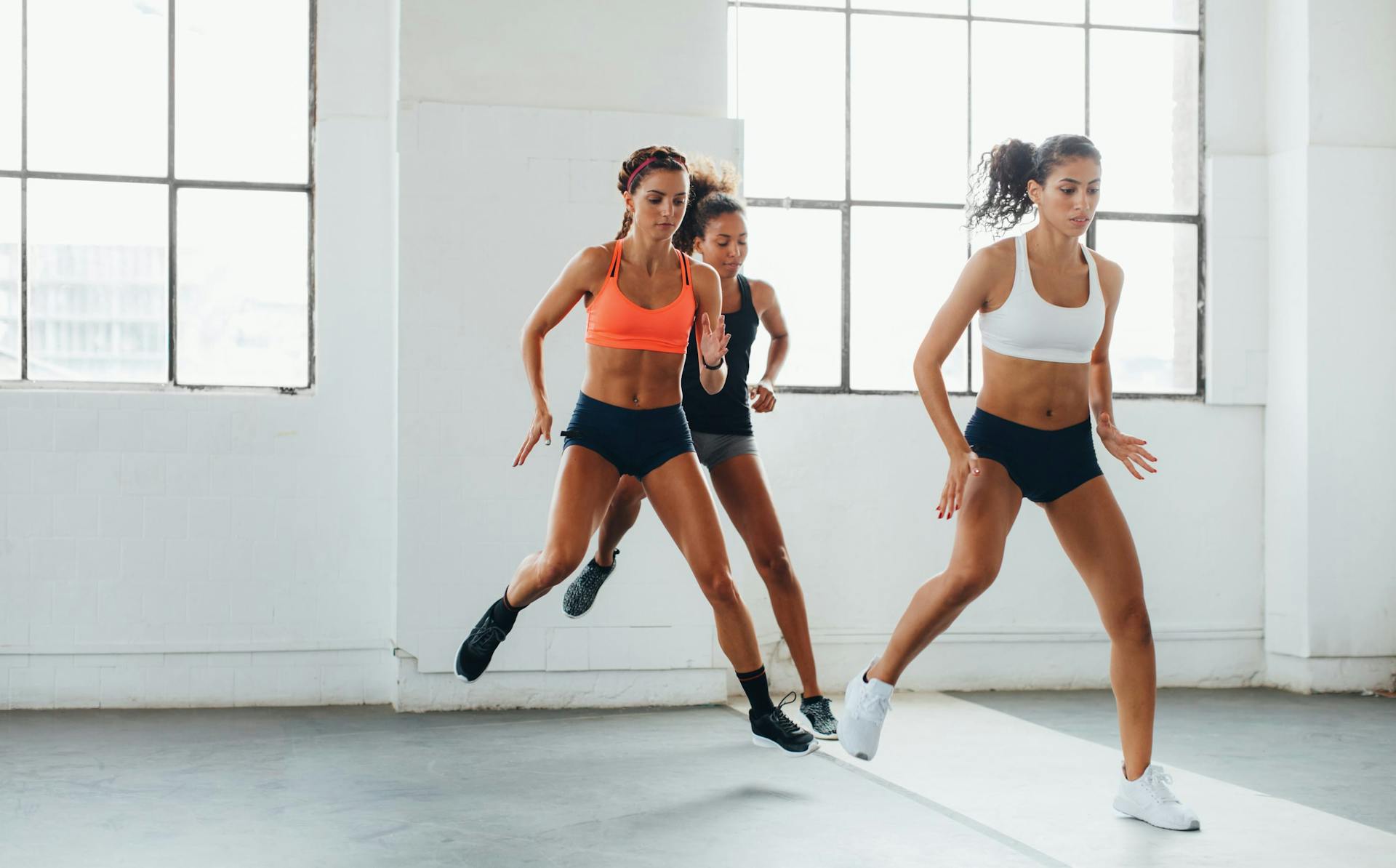 Tres mujeres con sujetadores deportivos y pantalones cortos en una clase de acondicionamiento físico en un gimnasio con paredes desnudas y grandes ventanales saltando de un pie a otro