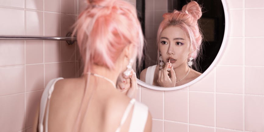 Una mujer asiática de cabello rosado se pone lápiz labial mientras se mira en el espejo de un baño rosa.