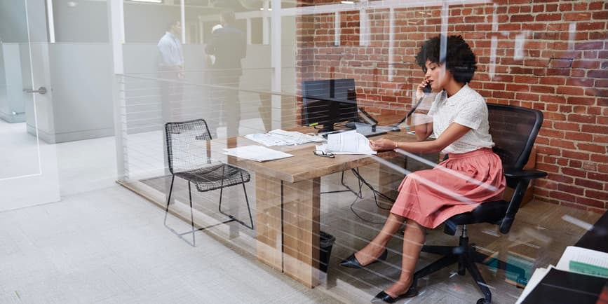 Color photograph through a glass wall looking into an office where a young, pretty Black woman with a short Afro wearing a white top and pink skirt looks down at paperwork while talking on the phone.