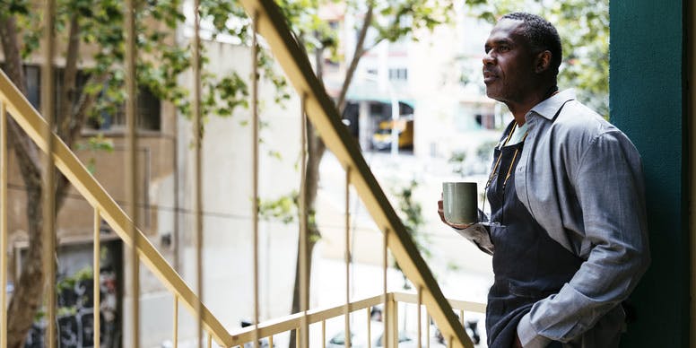 Vista lateral de un hombre negro con un delantal, de pie en las escaleras exteriores con una taza de café y mirando hacia otro lado de la cámara.