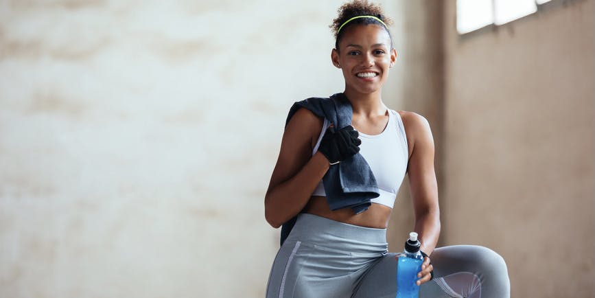A young Black woman with brown hair tied back wearing gym clothes smiles into the camera.