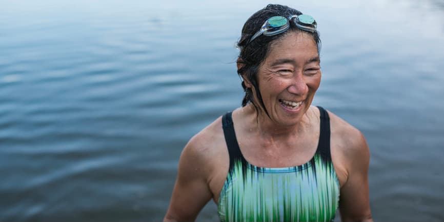 Retrato de una mujer bronceada de unos cincuenta o sesenta años que lleva un traje de baño y unas gafas, sonriendo al salir de un lago nadando.