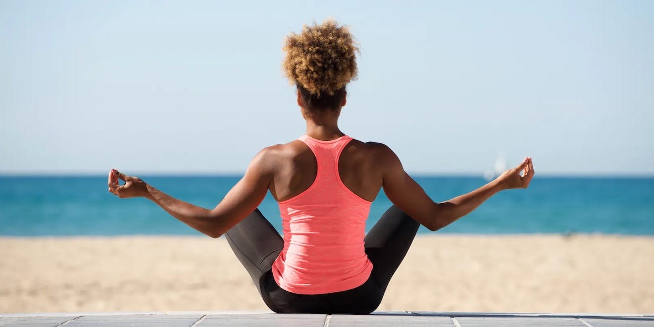 Una joven negra tonificada vestida con leggings negros y una camiseta sin mangas ajustada de color rosa brillante se sienta en la playa mirando hacia otro lado de la cámara mientras mantiene una postura de yoga elevada.