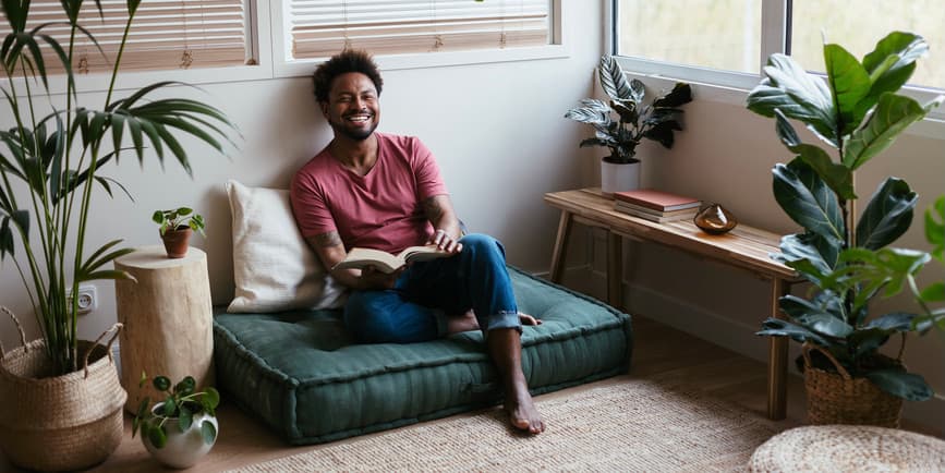 Fotografía de cuerpo entero de un joven negro vestido con ropa casual riéndose de un chiste mientras está sentado en un piso blando y leyendo un libro en su casa en una habitación iluminada por el sol, rodeada de plantas y detalles de madera y cuerdas.