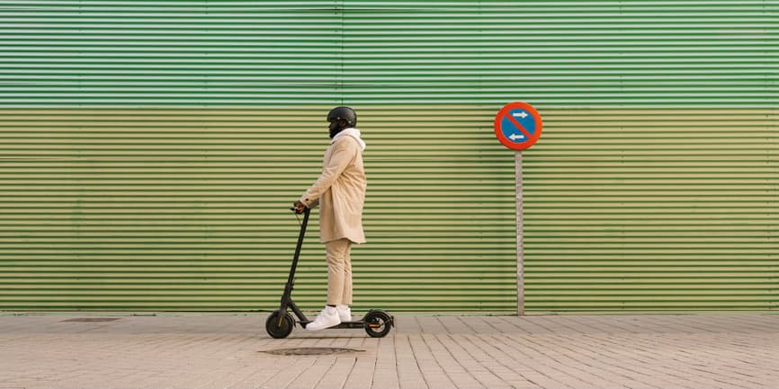 A Black man wearing a pale pink suit and helmet rides an electric scooter down a London sidewalk.