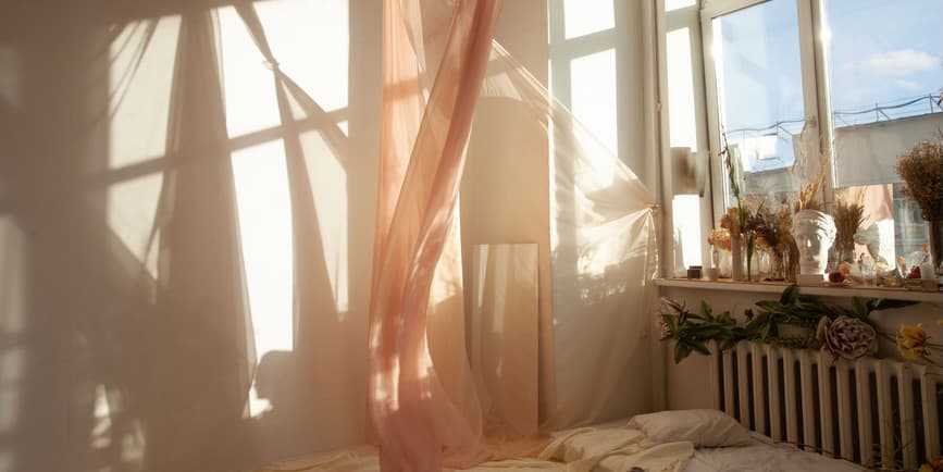 A dreamy indoor photograph of a bedroom where a gauzy curtain dances in the breeze of an open window and sunlight casts shadows on the wall.