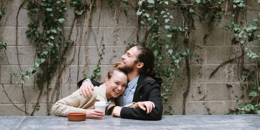 Pareja joven abrazándose y sonriendo mientras está sentada en el patio exterior de la cafetería