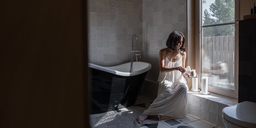 Una foto de ensueño de una joven blanca con un largo camisón blanco encendiendo velas en un baño con grandes azulejos a cuadros y una bañera con patas, con luz que entra por las ventanas.