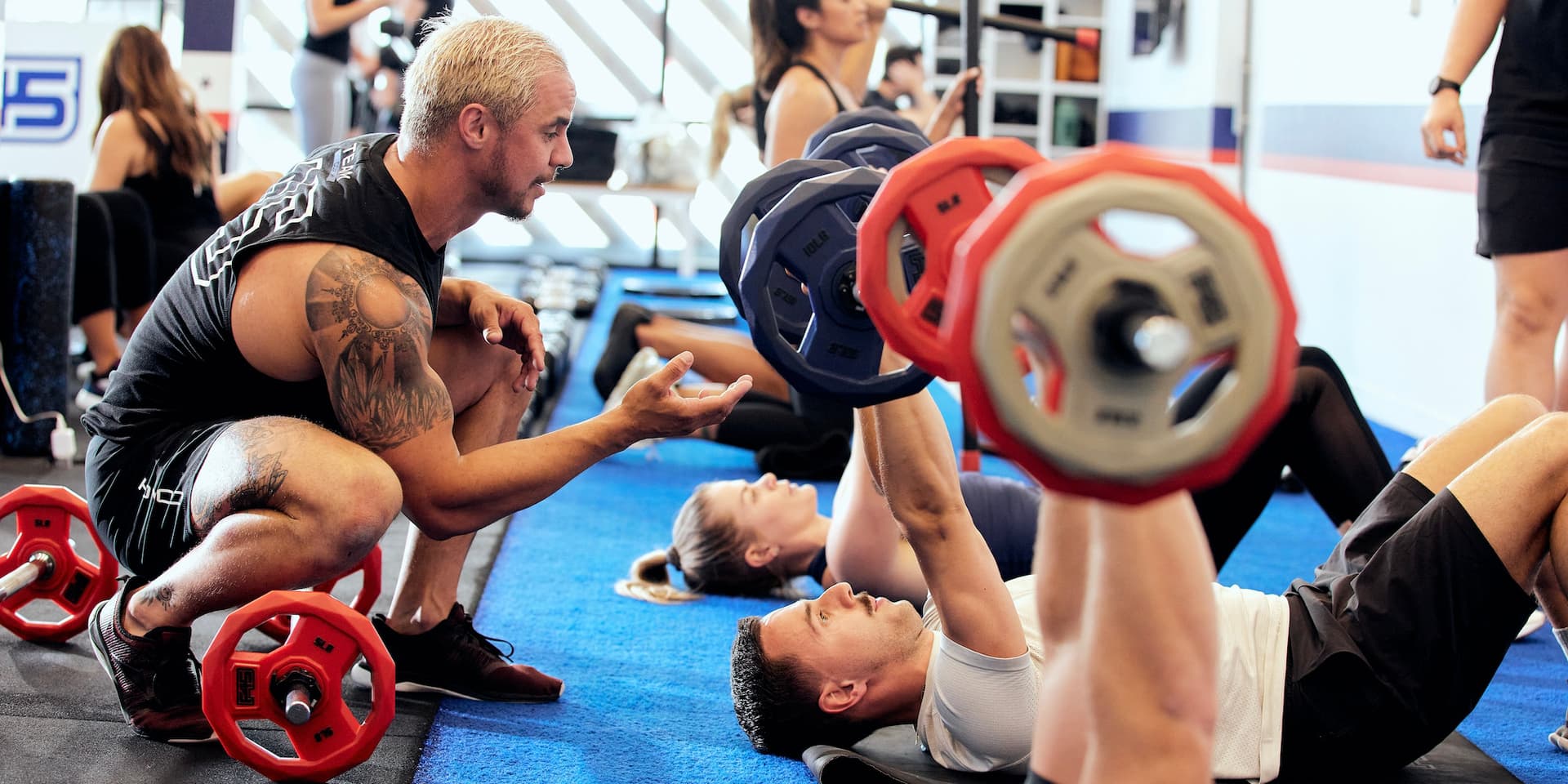 Fotografía en color de un entrenador personal masculino blanco de F45 Training arrodillado para ayudar a los miembros del gimnasio F45 que se tumban en el suelo, levantando pesas con barra por encima de ellos. Foto cortesía del F45.