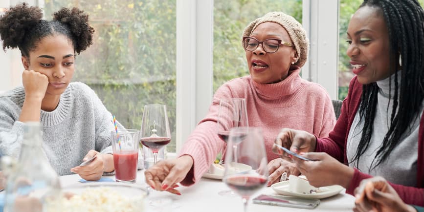 Smiling Black grandmother and her adult daughter and granddaughter sitting together at a table at home playing cards in a room with wide windows with greenery outside.