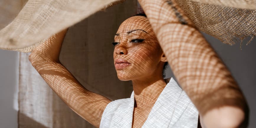 Mujer negra pensativa, maquillada y con los hombros descubiertos, que mira hacia otro lado mientras levanta una tela marrón a la luz del día y tiene puntos de luz en el cuerpo.