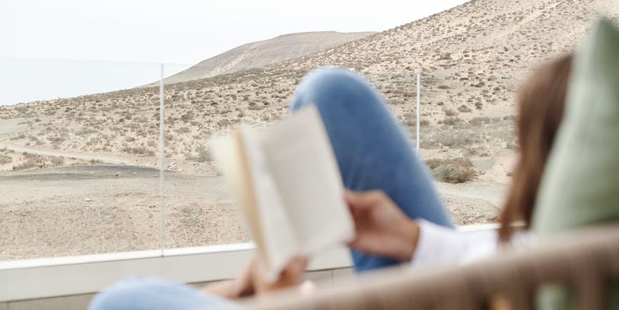 Mujer joven leyendo un libro mientras se relaja al aire libre en una silla en su patio con vistas al interior del Parque Nacional de Jandía con vistas al Océano Atlántico en la playa de Sotavento, Costa Calma, Fuerteventura, Islas Canarias, España.