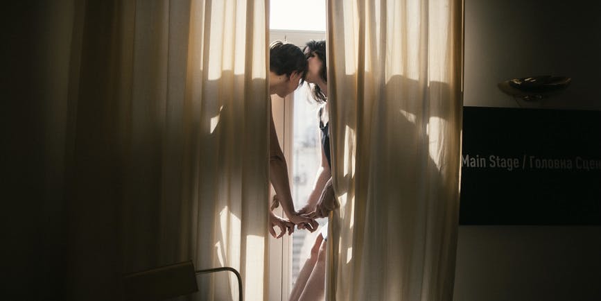 A young lesbian couple reaches toward each other behind gauzy curtains with light coming in from doors behind them.