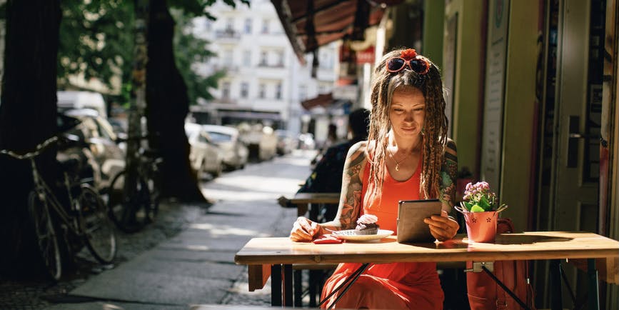 Una mujer blanca con largas rastas y tatuajes que lleva un vestido de verano se sienta en un café al aire libre, leyendo un libro electrónico.