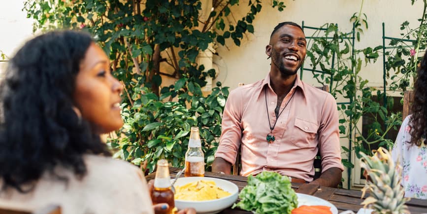 A multiethnic group of friends with a young handsome Black man wearing a pink shirt and smiling at center sits outside, eating and drinking on a patio at a table surrounded with greenery.