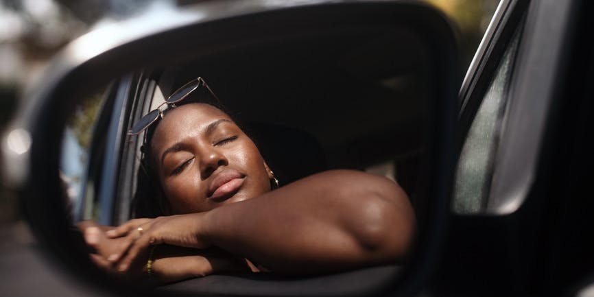 En el interior de un coche, una joven negra descansa con los ojos cerrados contra el sol y los brazos apoyados en el marco abierto de la ventana de la puerta, que se ve en el espejo lateral.