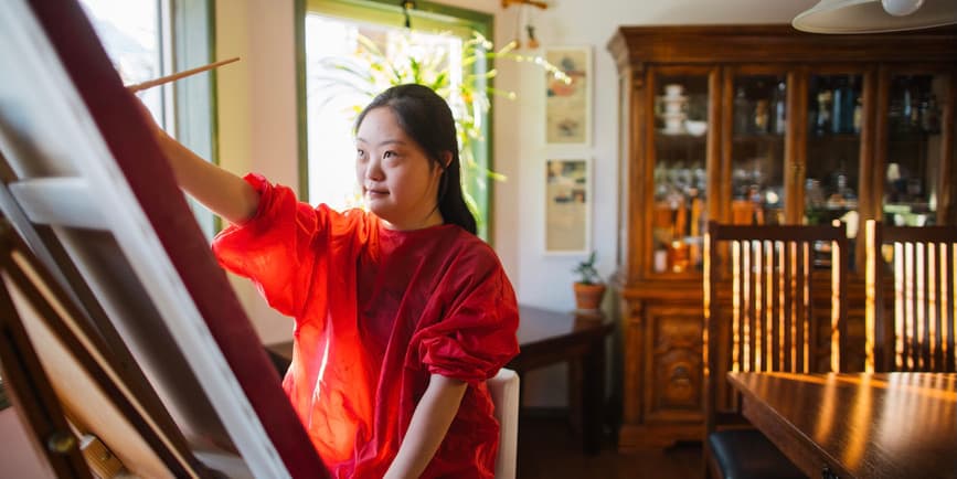 A young Asian woman with Downs syndrome wearing a red dress paints at an easel by windows in a brightly lit, warm dining room.