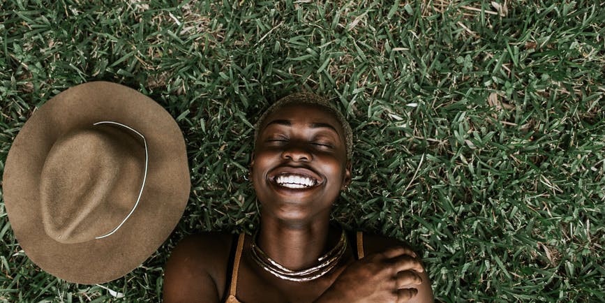 Retrato de una hermosa joven negra vestida con un top marrón sonriendo con los ojos cerrados mientras está tumbada en la hierba, con un sombrero marrón a su lado.