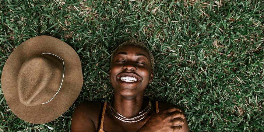 Retrato de una hermosa joven negra vestida con un top marrón sonriendo con los ojos cerrados mientras está tumbada en la hierba, con un sombrero marrón a su lado.