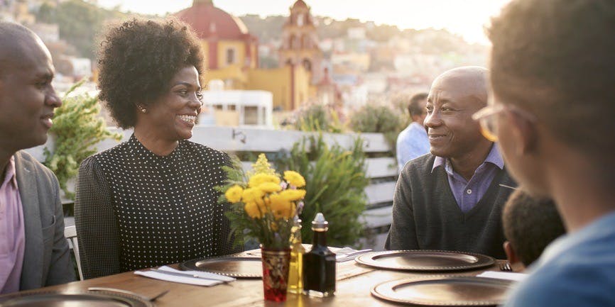 Tres generaciones de una familia negra sonriente cenan al aire libre al atardecer, en el restaurante de una ciudad montañosa y con una cálida iluminación.