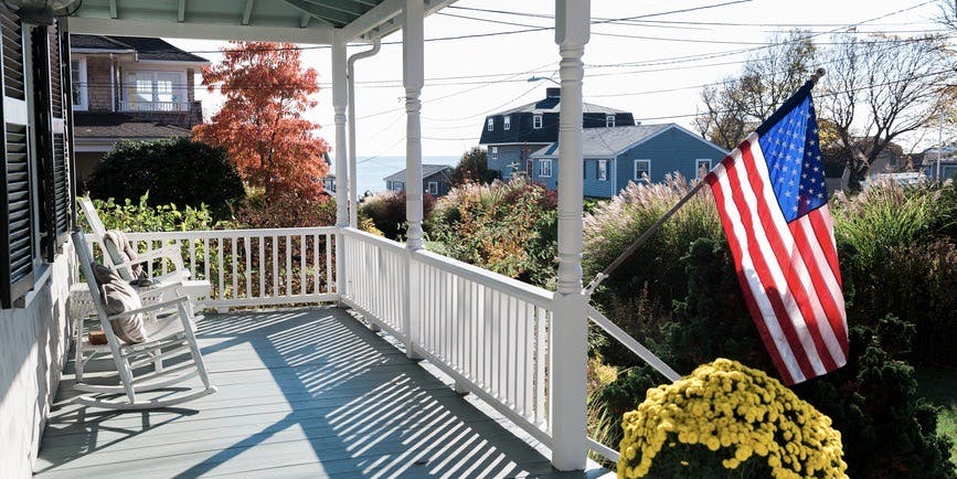 Un porche delantero con una valla blanca y dos mecedoras blancas, una bandera estadounidense ondeando con la brisa, los tejados de otras casas y el olor del agua a lo lejos.