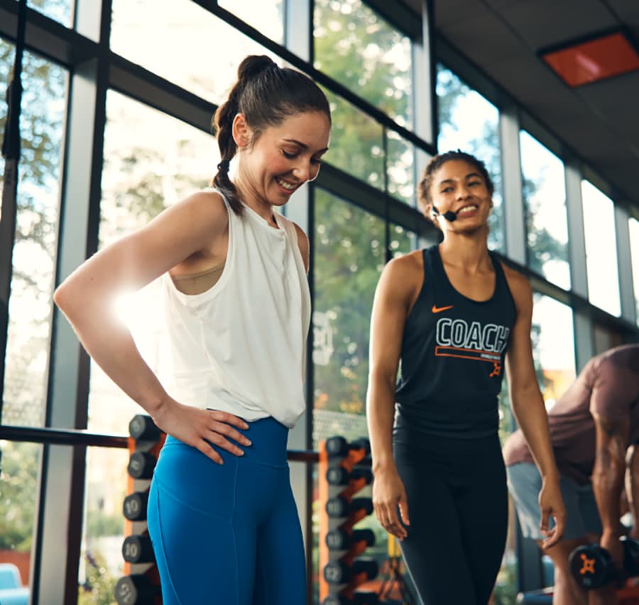 Two women are in a gym. One is wearing a white tank top and blue leggings, smiling. The other is a coach, wearing a black tank top with "COACH" printed on it.