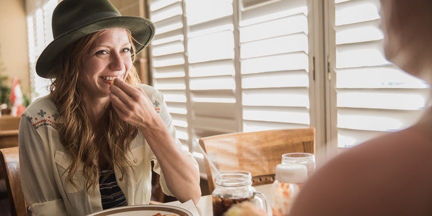 A young white woman with long wavy blond hair wearing a fedora and button down shirt smiles as she eats a french fry while sitting at a diner booth next to a window with sun streaming through.