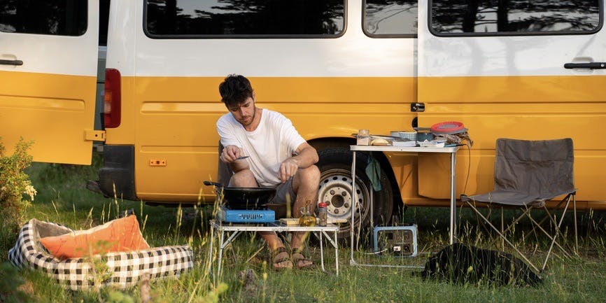 An outdoor summery photo of a young white man sitting in front of a yellow and white camper van, making food at a camping stove with a table of equipment nearby and a dog bed and black dog lying in the grass.