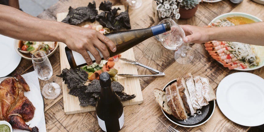 An overhead photo of a brown arm pouring wine above a table set with colorful, healthy food and the suggestion of friends sitting and enjoying a meal together.