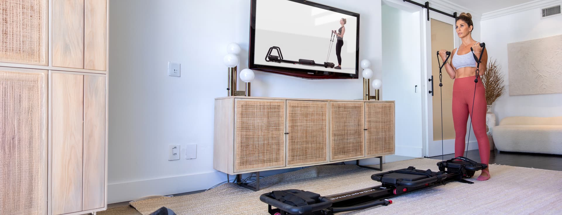 A woman in a home setting exercising with resistance bands attached to a reformer machine, while following along with a workout video on a wall-mounted TV.