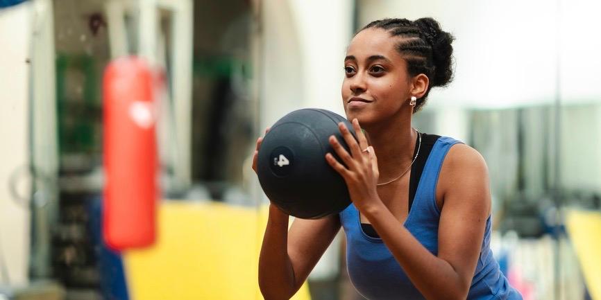 Determinada atleta etíope con un moño de pelo trenzado que se pone en cuclillas con una pelota medicinal pesada durante un intenso entrenamiento en el gimnasio.