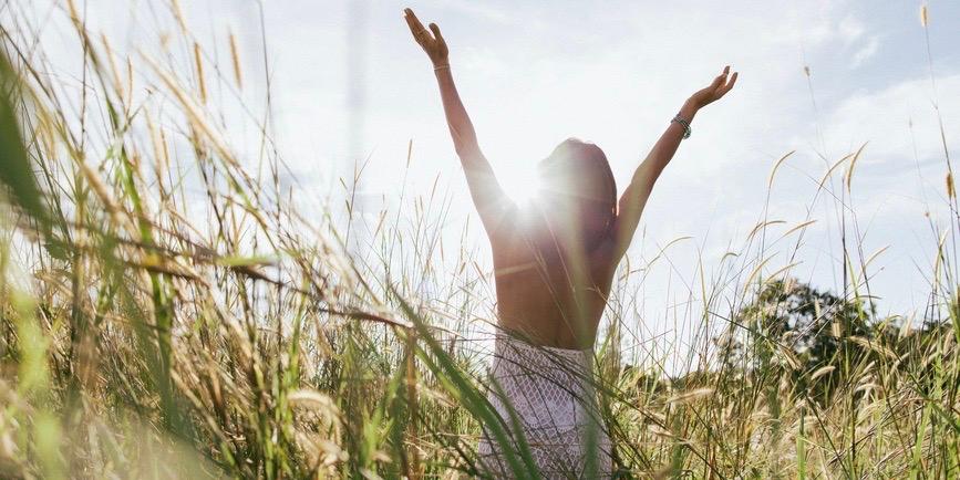 La espalda de una mujer joven de pie en un campo de flores silvestres durante la puesta de sol, con los brazos levantados.