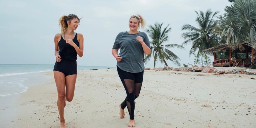 Dos jóvenes blancas, ambas de cabello rubio, una delgada y otra con curvas, corren por una playa de Tailandia, sonriendo y riendo juntas.