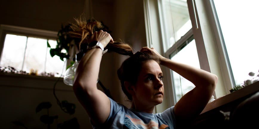 A shadowy portrait of a white woman sitting in front of a window looking out while putting her hair into a ponytail.