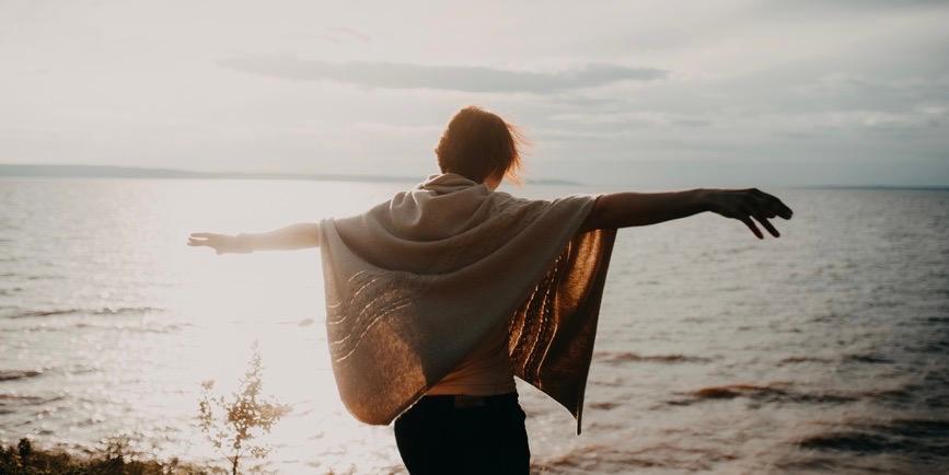 Retrato desde atrás de una mujer joven en la playa levantando las manos hacia el sol.