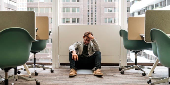 Worried guy in casual wear thinking about problem sitting on floor near with closed laptop touching head in office.
