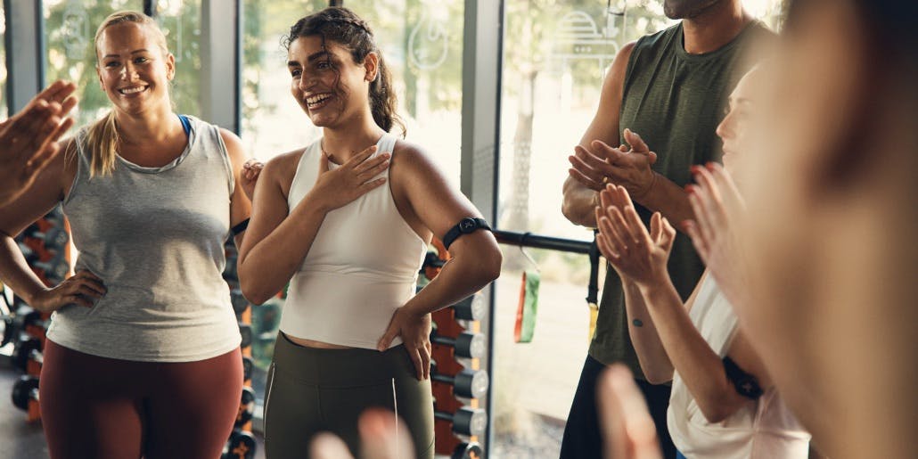 A young brown-skinned woman with brown hair smiles as she's being applauded by Orangetheory Fitness classmates.