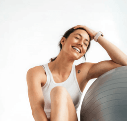 Una mujer con ropa deportiva sentada y sonriendo apoyada en una pelota de entrenamiento