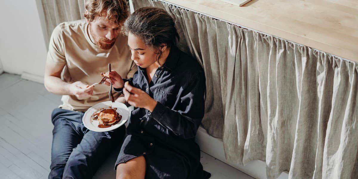 A young white man with red hair and young Latina woman with dark hair share a plate of pancakes while sitting on their kitchen floor.