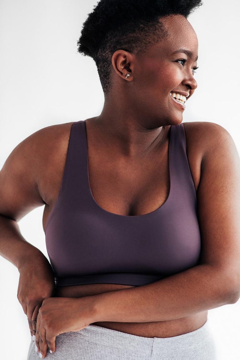 Frente a un fondo neutro, mujer vestida con ropa deportiva sujetándose la parte derecha de la cintura con ambas manos, mirando y sonriendo hacia la izquierda.