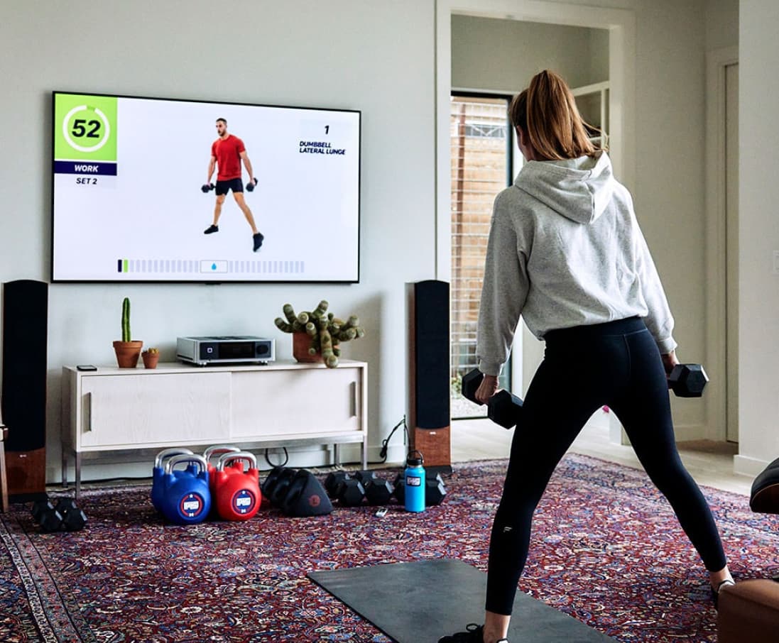 Mujer en casa con pesas de entrenamiento haciendo ejercicio frente a un televisor siguiendo un programa de entrenamiento de F45.