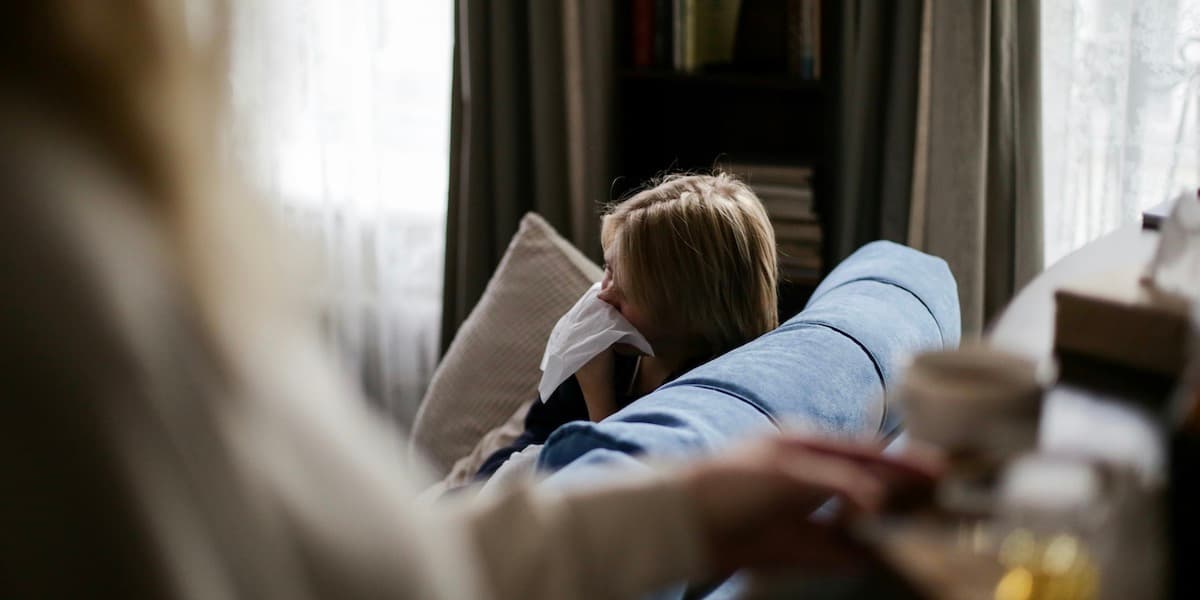 A person with blond hair and their face obscured by a tissue sits on a blue couch, while someone closer to the camera prepares a cup of tea with honey.