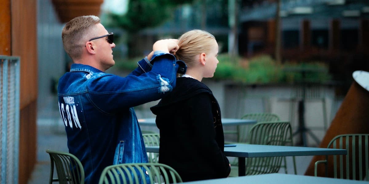 A white father with blond hair wearing a blue jersey sits outside at otherwise empty metal tables, braiding the blond hair of his young daughter.